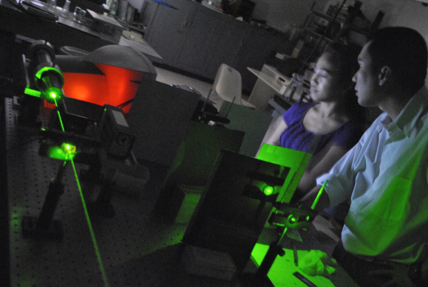A quantum optics laboratory: green laser beams steered across an optical table through mirrors and lenses while two researchers look on